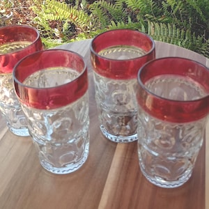 May include: Four clear drinking glasses with a red rim, arranged on a wooden surface. The glasses have a textured pattern on the sides. The background includes green foliage, suggesting an outdoor setting.