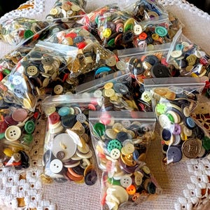 May include: A collection of clear plastic bags filled with assorted sewing buttons in various shapes, sizes, and colors, including red, blue, green, yellow, black, white, and metallic tones, featuring different hole counts and textures.
