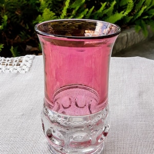 May include: A vintage, clear glass tumbler with a vibrant pink hue. The upper portion of the glass is a solid pink, while the lower section features a detailed, embossed design. The glass is set against a neutral background with green foliage in the background.