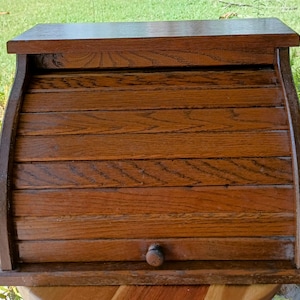 May include: A wooden breadbox with a roll-top door. The breadbox is made of dark brown wood with a visible wood grain. It has a rounded top and a small wooden knob on the front. The breadbox is sitting on a wooden surface.