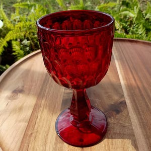 May include: A ruby red vintage goblet with a textured pattern. The glass has a round base, a stem, and a detailed bowl. The goblet is sitting on a wooden surface, with green foliage in the background.