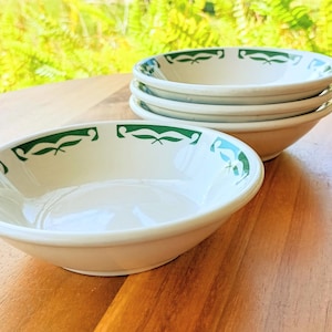 May include: A stack of four off-white ceramic bowls with a green leaf design around the rim. One bowl is in the foreground. The bowls are on a wooden surface with a blurred green background.