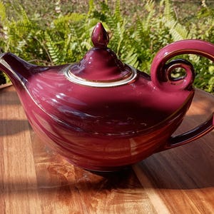 May include: A burgundy ceramic teapot with a gold trim and a decorative handle. The teapot has a curved spout and a matching lid with a finial. The teapot is sitting on a wooden surface.