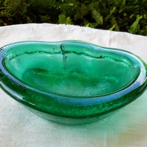 May include: A green glass bowl with a wavy, organic shape. The bowl is made of thick glass and has a smooth, polished finish.