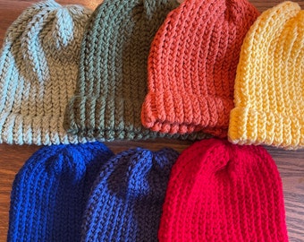 May include: A collection of seven knitted beanies in various colors, including sage green, olive green, orange, yellow, blue, and red. The beanies are arranged in a close-up shot, showcasing their ribbed texture and folded brims. The hats are made of a soft, warm material.