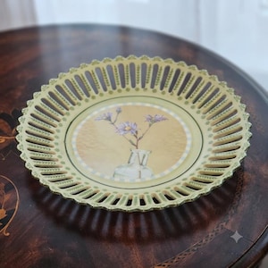 May include: A round ceramic plate with a decorative openwork lattice rim in pale yellow and green. The center features a circular illustration of purple flowers in a clear glass vase set against a soft, warm, light-brown background.