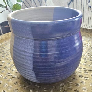 May include: A round ceramic planter featuring a two-tone glaze design. One side of the pot is finished in a light cream color, while the other side is a deep, vibrant blue. The exterior surface displays subtle horizontal ridges throughout the body.