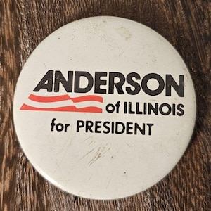 May include: A white campaign button with the text "ANDERSON of ILLINOIS for PRESIDENT" in black lettering. The button features a red and white striped design above the words "of ILLINOIS". The button is round and has a slightly worn appearance.