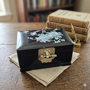 May include: A rectangular black lacquered jewelry box featuring an iridescent mother-of-pearl inlay of flowers and a bird on the lid. The box includes a decorative brass latch shaped like a fish, providing a secure closure for the storage container.