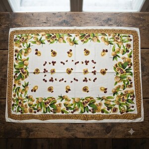 May include: A rectangular white fabric cloth featuring a border of brown woven rope patterns. The surface is decorated with a repeating botanical print of green leaves, spiky yellow chestnut husks, and dark brown chestnuts scattered throughout.