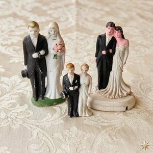 May include: A collection of vintage wedding cake toppers. The figurines depict a bride and groom in various poses, with the grooms in black tuxedos and the brides in white gowns. Some brides hold bouquets. The toppers are on a cream-colored surface.