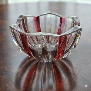 May include: A clear glass bowl with a red stripe design. The bowl has a unique, geometric shape with a star-like rim. The bowl is sitting on a wooden surface.