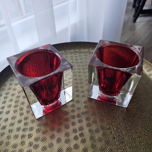 May include: Two square glass candle holders featuring a clear outer cube frame and a deep red, rounded interior vessel. The design combines geometric transparency with a vibrant, reflective crimson color, creating a distinct contrast between the outer edges and the inner bowl shape.