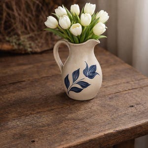 May include: A beige ceramic pitcher with a dark blue floral design holds a bouquet of white tulips. The pitcher has a handle and is set on a rustic wooden table. The flowers are in full bloom, creating a fresh, natural aesthetic.