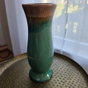 May include: A tall ceramic vase with a flared rim and a pedestal base. The body features a smooth, glossy finish with a crackle glaze texture, transitioning from a deep teal green at the bottom to a warm, mottled brown at the top rim.