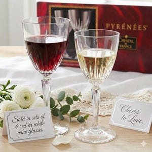 May include: Two crystal wine glasses, one filled with red wine and the other with white wine, are displayed. The glasses are in front of a box that says "PYRÉNÉES CRYSTAL FRANCE". A card reads "Sold in sets of 4 red or white wine glasses."