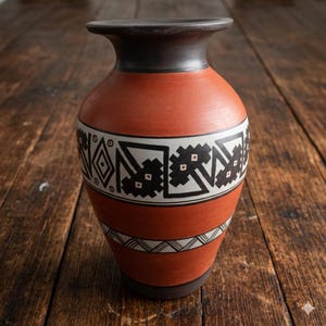 May include: A terracotta-colored clay vase with a dark gray rim and base. The body features a wide band of black and white geometric patterns, including diamonds and jagged shapes, and a thinner lower band with a repeating chevron design.