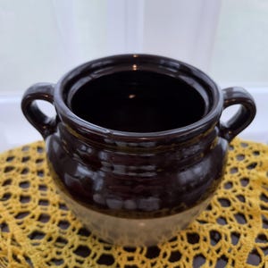 May include: A two-handled ceramic pot featuring a glossy dark brown glaze on the upper portion and a smooth, lighter beige finish on the lower section. The vessel has a wide, open circular mouth and two curved side handles for easy gripping.