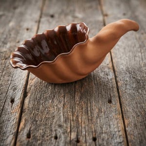 May include: A ceramic bowl shaped like a seashell with a matte terracotta exterior and a glossy dark brown interior. The bowl features a curved, elongated handle extending from one side, with a scalloped rim and a fluted, ribbed texture.