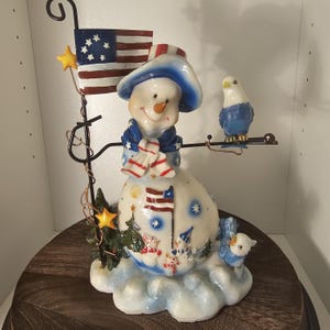May include: A decorative snowman figurine featuring a blue hat, a scarf with red and white stripes, and a body painted with American flags and small snowmen. A metal pole holds a flag and a blue bird, with a second blue bird perched at the base.