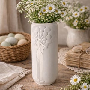 May include: A white ceramic vase with a tree-like embossed design, filled with white daisies and greenery. The vase is on a wooden surface, with a basket of eggs and a stack of books in the background.