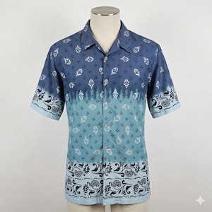 May include: A short-sleeved button-down shirt in shades of blue, featuring a gradient design. The top portion has a dark blue pattern, transitioning to a lighter blue with a paisley border at the bottom. The shirt is displayed on a mannequin.