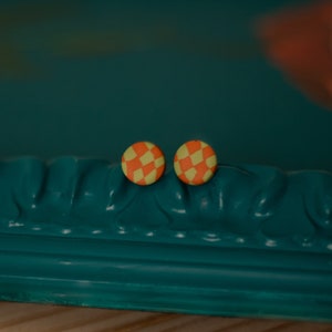 Puede incluir: Un par de pendientes de botón a cuadros naranja y amarillo sobre una superficie verde azulada.