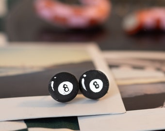 May include: A pair of black 8-ball earrings. The earrings are made of polymer clay and are shaped like a pool ball. The earrings are on a white surface.