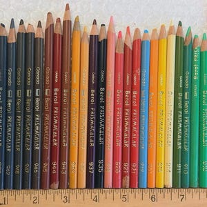 May include: A collection of Berol Prismacolor pencils in various colors, arranged vertically. The pencils are labeled with numbers and the brand name "Berol Prismacolor" and "Canada". A wooden ruler is placed next to the pencils.