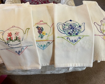 May include: A set of white tea towels, each embroidered with a different teapot design. The teapots are adorned with colorful floral patterns and sit atop a decorative embroidered base. The towels are made of a soft, absorbent material.