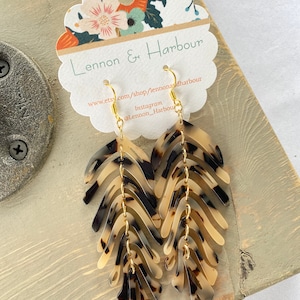 Puede incluir: Un par de aretes largos y colgantes con un patrón de caparazón de tortuga. Los aretes están hechos de múltiples capas de plástico, cada una con una forma diferente. Los aretes están conectados por anillos de metal dorado. Los aretes cuelgan de una tarjeta blanca con el texto "Lennon & Harbour" y una dirección de sitio web.