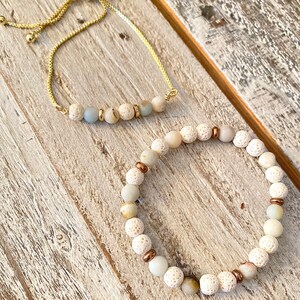 May include: Two bracelets with white and brown beads. One bracelet is a gold chain with a slider clasp and the other is a beaded bracelet with white lava rock beads and gold accents.