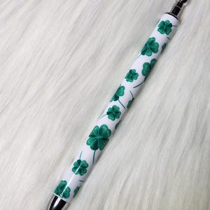 May include: A white pen with a pattern of green shamrocks. The pen has a silver tip and a silver stylus on the opposite end. The shamrocks are in various sizes and are evenly spaced across the pen's surface.