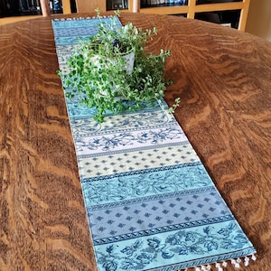 May include: A patterned table runner in shades of blue, green, and cream, with a beaded fringe. The runner features floral and geometric designs. A potted plant rests on the runner, which is placed on a wooden table. The runner is approximately 150 cm long.