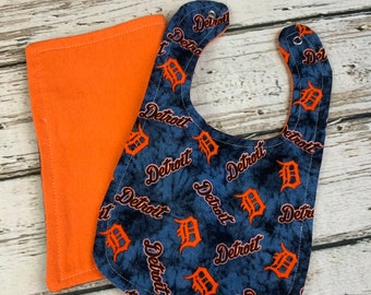 May include: A blue and orange baby bib with a tie-dye pattern and the word "Detroit" repeated throughout. The bib is paired with a solid orange burp cloth.