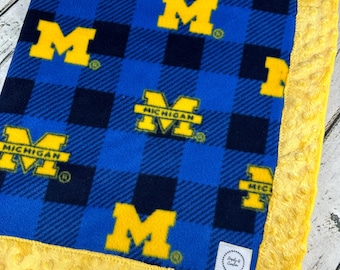 May include: A blue and yellow fleece blanket with a plaid pattern. The blanket features the letter "M" and the word "MICHIGAN" in yellow. The border is a solid yellow with a textured, dotted pattern. A small white tag is in the corner.