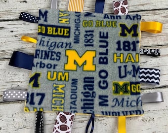 Puede incluir: Una manta para bebé de color gris y amarillo con varios lazos atados. La manta presenta el logotipo de la Universidad de Michigan y las palabras "Go Blue" y "Michigan".