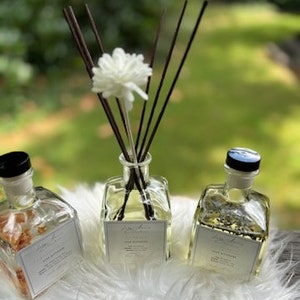 K&ouml;nnte beinhalten: Drei transparente Aromatherapie-Diffusorflaschen mit schwarzen Kappen und wei&szlig;en Etiketten. Eine Flasche hat eine wei&szlig;e Blume und dunkelbraune Schilfrohre. Die anderen beiden Flaschen haben eine hellgelbe Fl&uuml;ssigkeit. Die Flaschen stehen auf einer wei&szlig;en, flauschigen Oberfl&auml;che.