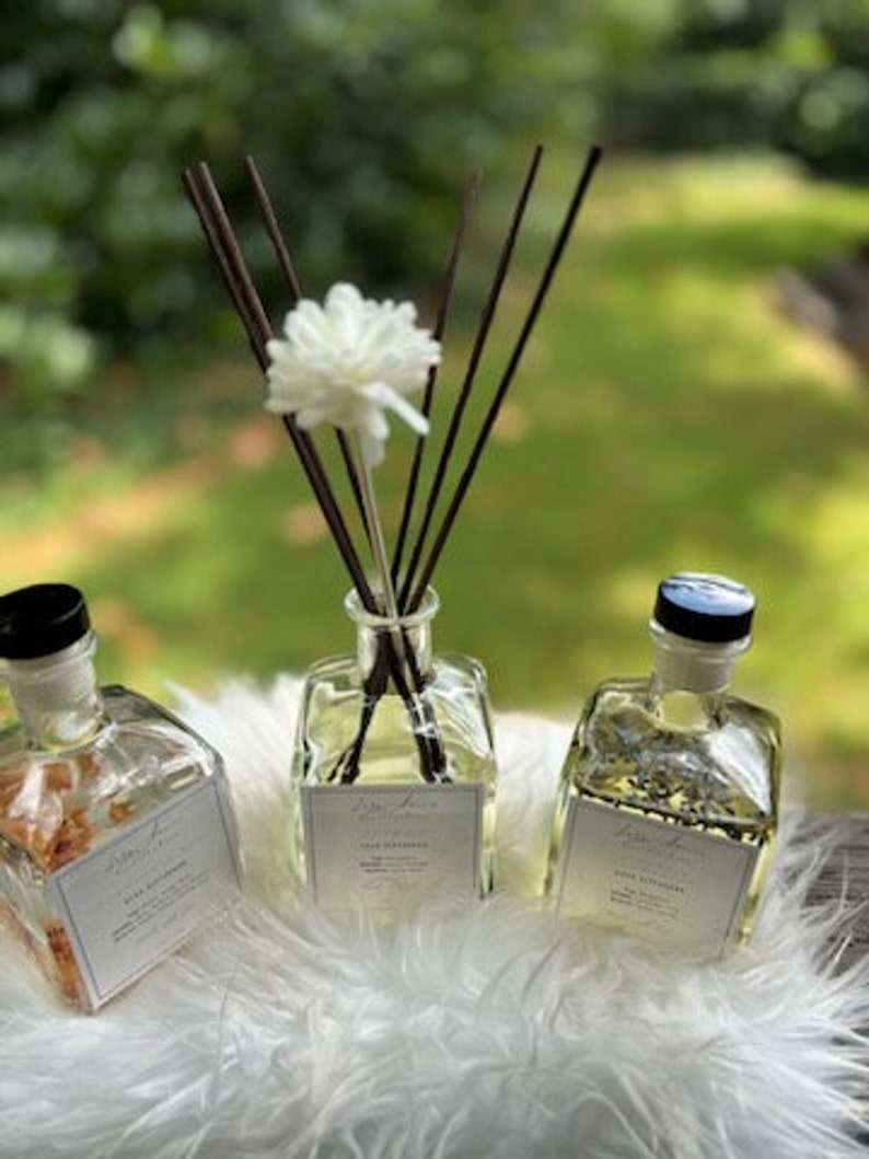 K&ouml;nnte beinhalten: Drei transparente Aromatherapie-Diffusorflaschen mit schwarzen Kappen und wei&szlig;en Etiketten. Eine Flasche hat eine wei&szlig;e Blume und dunkelbraune Schilfrohre. Die anderen beiden Flaschen haben eine hellgelbe Fl&uuml;ssigkeit. Die Flaschen stehen auf einer wei&szlig;en, flauschigen Oberfl&auml;che.