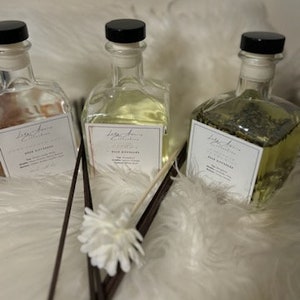 K&ouml;nnte beinhalten: Drei Glas-Diffusorflaschen mit schwarzen Deckeln und wei&szlig;en Etiketten. Die Flaschen enthalten unterschiedlich farbige Fl&uuml;ssigkeiten und Pflanzenstoffe. Dunkelbraune Diffusorst&auml;bchen und eine wei&szlig;e Blume befinden sich vor den Flaschen.