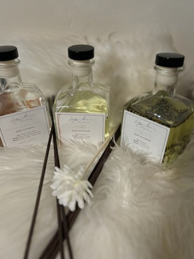 K&ouml;nnte beinhalten: Drei Glas-Diffusorflaschen mit schwarzen Deckeln und wei&szlig;en Etiketten. Die Flaschen enthalten unterschiedlich farbige Fl&uuml;ssigkeiten und Pflanzenstoffe. Dunkelbraune Diffusorst&auml;bchen und eine wei&szlig;e Blume befinden sich vor den Flaschen.