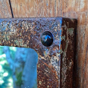 Puede incluir: Primer plano de un espejo rectangular con un marco de metal muy oxidado. El marco tiene un aspecto oscuro y texturizado con remaches visibles. El espejo refleja una escena exterior borrosa.