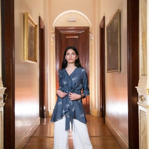 May include: A woman wearing a blue patterned blouse with a white tie and white wide-leg pants. She is standing in a hallway with paintings on the walls.