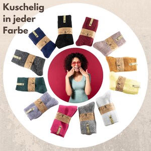 May include: A selection of colourful socks arranged in a circle around a woman wearing sunglasses. The socks are all made from a soft, cosy material and come in a range of colours, including grey, black, red, pink, yellow, and blue. The text "Kuschelig in jeder Farbe" is written at the top of the image.