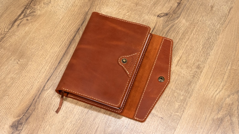 May include: A brown leather journal with a flap closure and a small pocket on the front. The journal is open to reveal a blank page.
