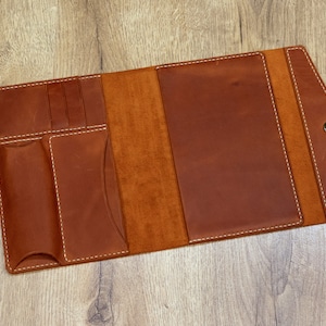 May include: A brown leather notebook with a flap closure and multiple pockets. The notebook is open to reveal a blank interior.