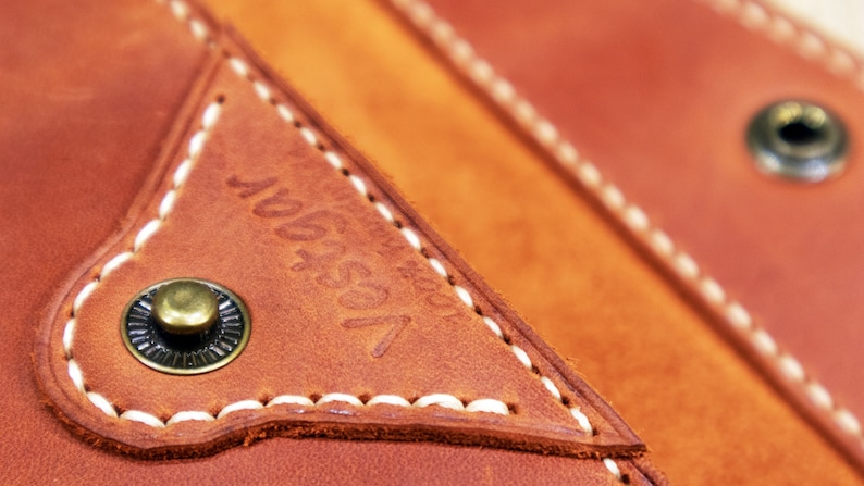 May include: Close-up of a brown leather bag with white stitching and a brass button. The bag has a triangular pocket with the text "1954" embossed on it.