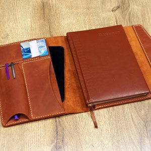 May include: A brown leather notebook cover with a built-in pocket for a phone or tablet, two pen holders, and a card slot. The cover is open to reveal a brown leather-bound notebook with the word "Planner" embossed on the front.