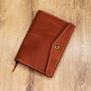May include: A brown leather journal with a flap closure and a brass button. The journal has a bookmark ribbon and is lying on a wooden surface.