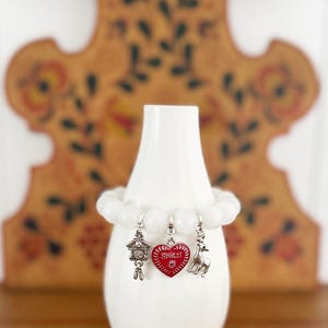 Op de afbeelding: Een armband van gladde, ronde, ondoorzichtige witte kralen op een elastisch koord. De armband is voorzien van drie zilverkleurige bedels: een koekoeksklok, een rood hartvormig bedeltje met de tekst Spatzl en een klein hertfiguurtje.